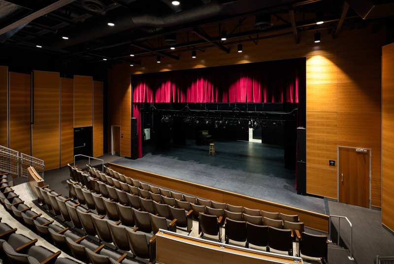The interior of the DeLuxe Theater depicting a stage. The walls are of wood panels and a red velvet curtain.
